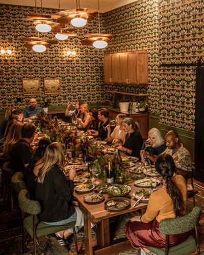Group of about 14 people dining around a long wooden table in a warmly lit restaurant with patterned wallpaper.
