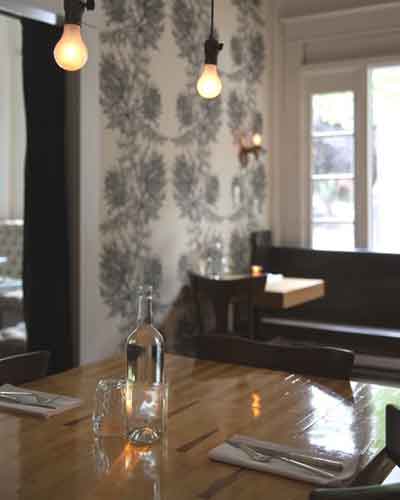 Restaurant dining table with a water bottle, glass, and cutlery under warm pendant lights, patterned wallpaper in the background.