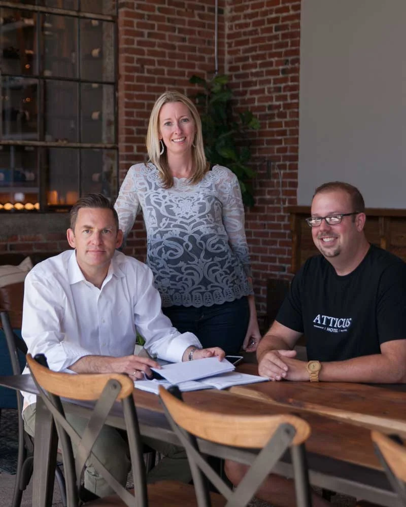 The Atticus Hotel ownership team at a wooden table with documents in a brick-walled office setting.