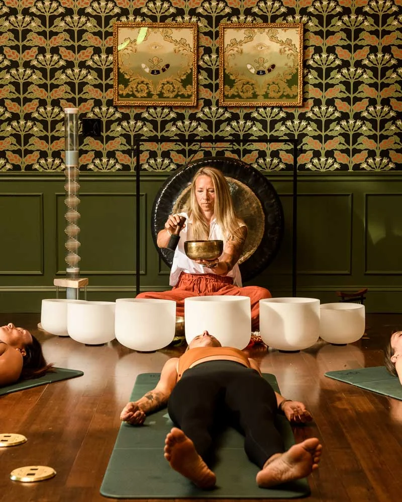 Meditation room with a teacher playing a singing bowl while students lie on yoga mats.