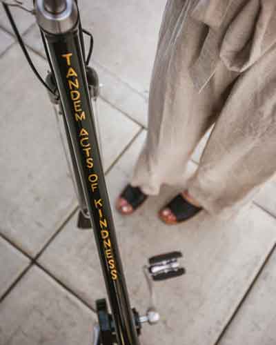 Close-up of a scooter frame with orange vertical text 'TANDEM ACTS OF KINDNESS'; a person in beige pants and sandals stands on a tiled surface, with two small objects on the ground nearby.