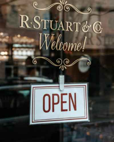 Storefront window with decorative lettering reading 'R. Stuart & Co Welcome!' and a hanging 'OPEN' sign in the foreground.