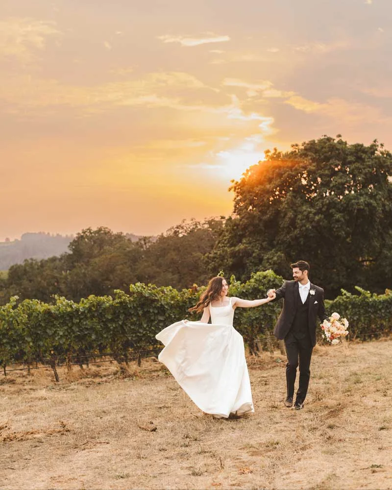 Bride in a white wedding gown twirls as a groom in a dark suit walks beside her in a vineyard at sunset.