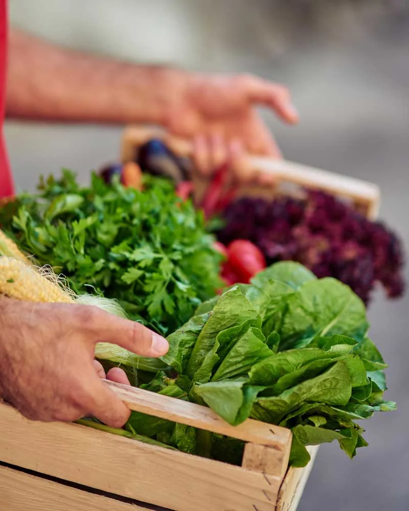 Person handling a wooden crate full of fresh leafy greens at a market stall, with other vegetables in the background