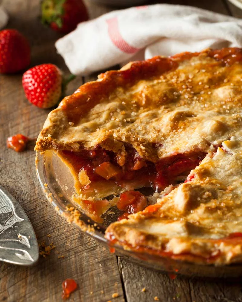 Strawberry pie with a flaky golden crust on a rustic wooden table, a slice pulled out to reveal red strawberry filling.