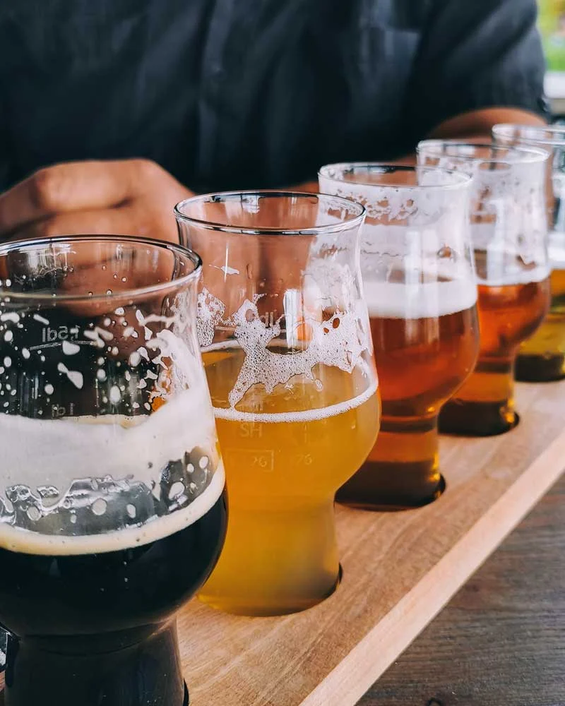Row of beer glasses with varying colors and foam on a wooden flight board, viewed from the side.