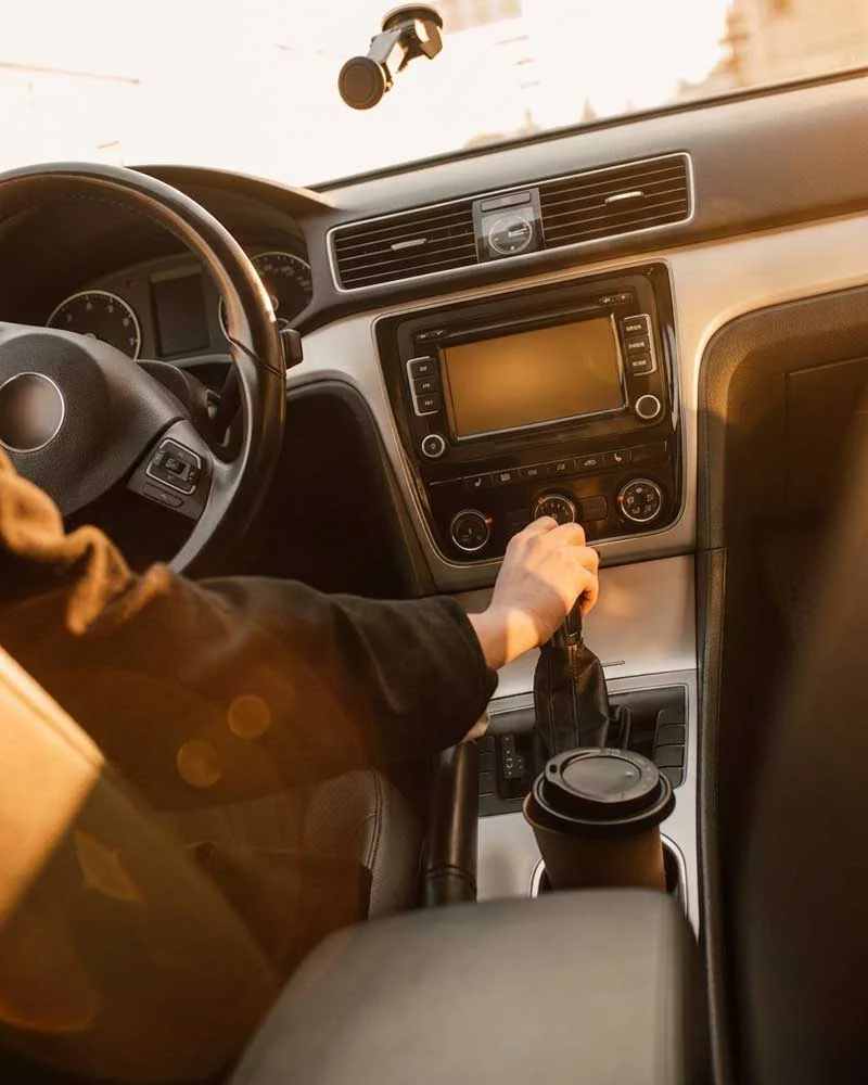 Driver shifts gears in a car's center console while a coffee cup sits in the cup holder near the passenger seat.