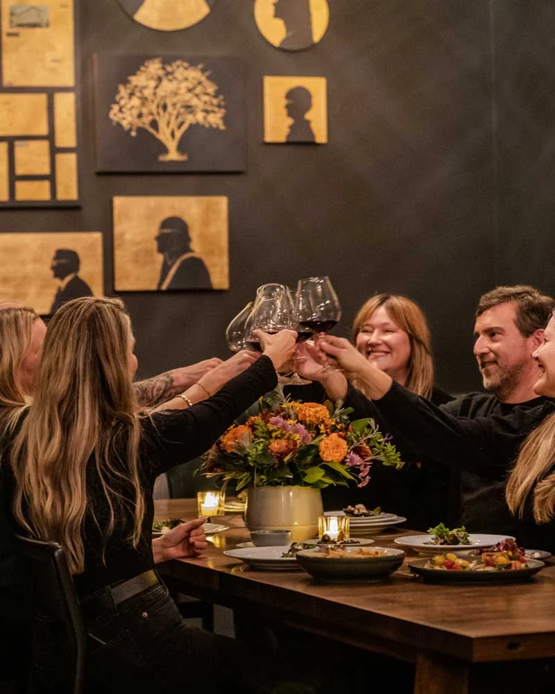 Group of friends at a dinner party raising wine glasses in a toast, with a floral centerpiece on the table.