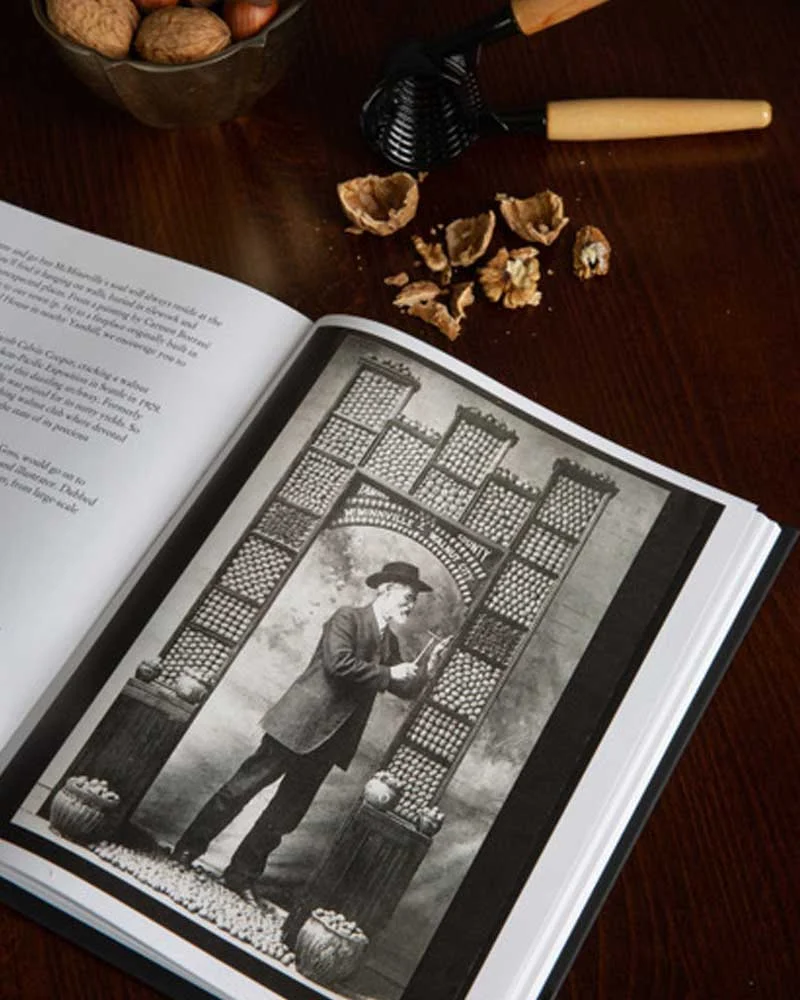 Attiicus-Hotel-Philosophy-Cooper Open book displaying a vintage photo of a man in a suit and hat beside a lattice arch, with a nutcracker and walnut shells on the wooden table.