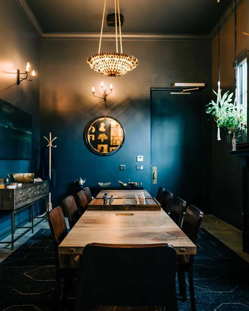Attiicus-Hotel-Philosophy-Our-Values Long wooden dining table with a chessboard centerpiece in a dark blue-walled room, with a chandelier hanging above.