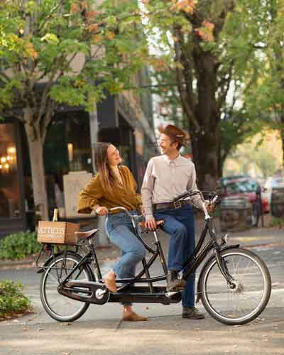 McMinnville-downtown-on-tandem-bikes Two people ride a tandem bicycle on a tree-lined city street, smiling at each other.